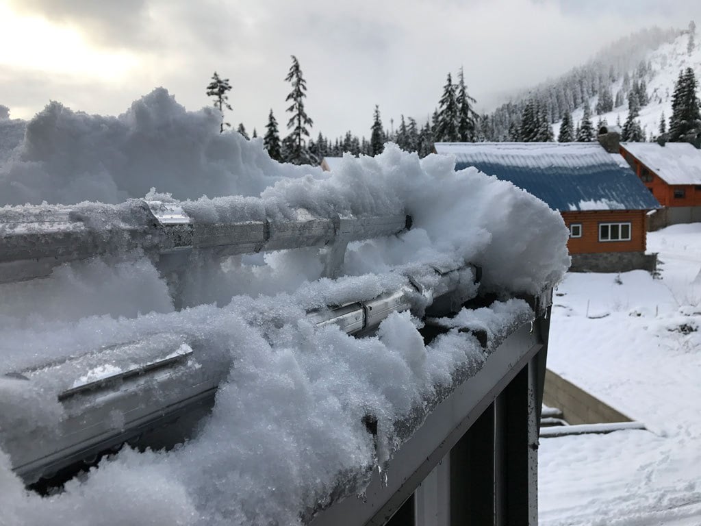 A metal gutter heavily covered in thick snow, with a blue-roofed building and snowy mountains in the background.