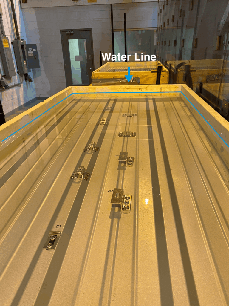 Línea de agua visible en una mesa grande, con soportes S-5! instalados en una muestra de material de techo de metal sumergido en agua.