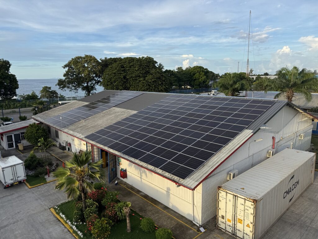Almacén con paneles solares en el tejado cerca del océano, rodeado de palmeras, vegetación, contenedores de transporte y un camión de reparto.