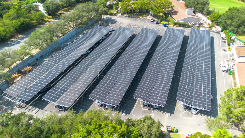 Vista aérea de un sistema de cocheras solares con múltiples filas de estructuras inclinadas cubiertas con paneles fotovoltaicos sobre un estacionamiento.