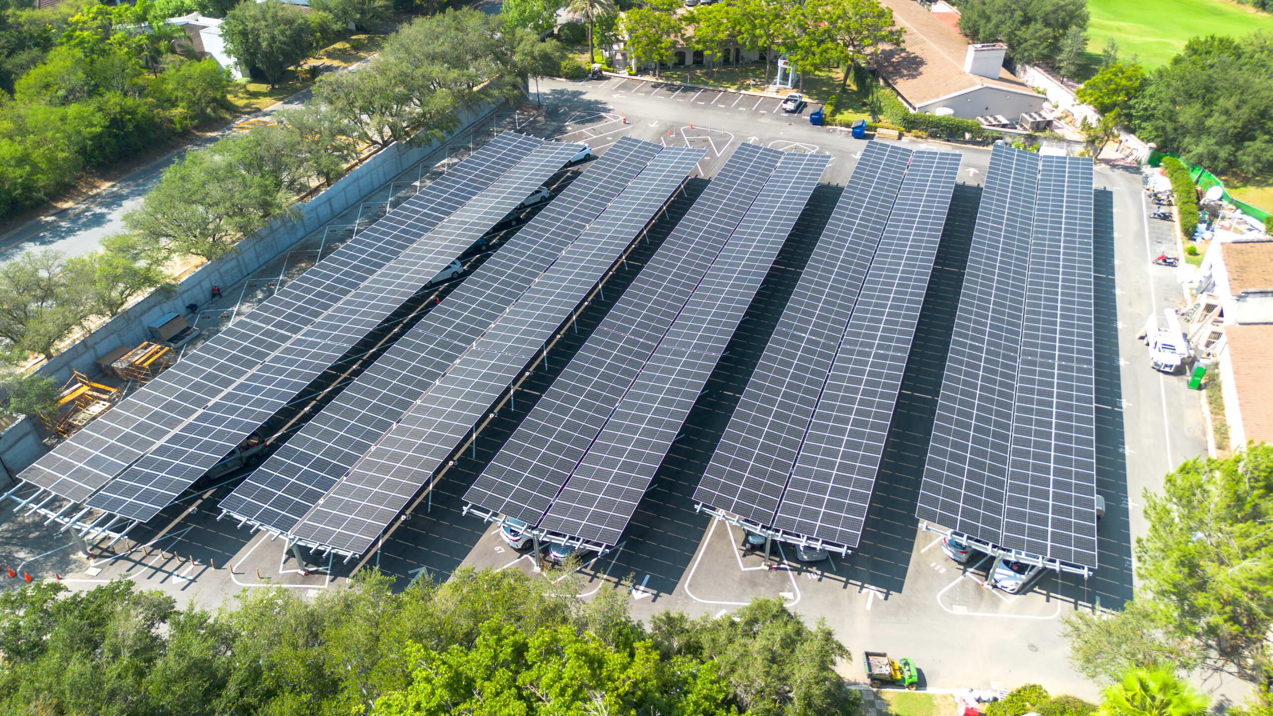 Aerial view of solar panel carports covering rows of parking spaces in a lot surrounded by trees.