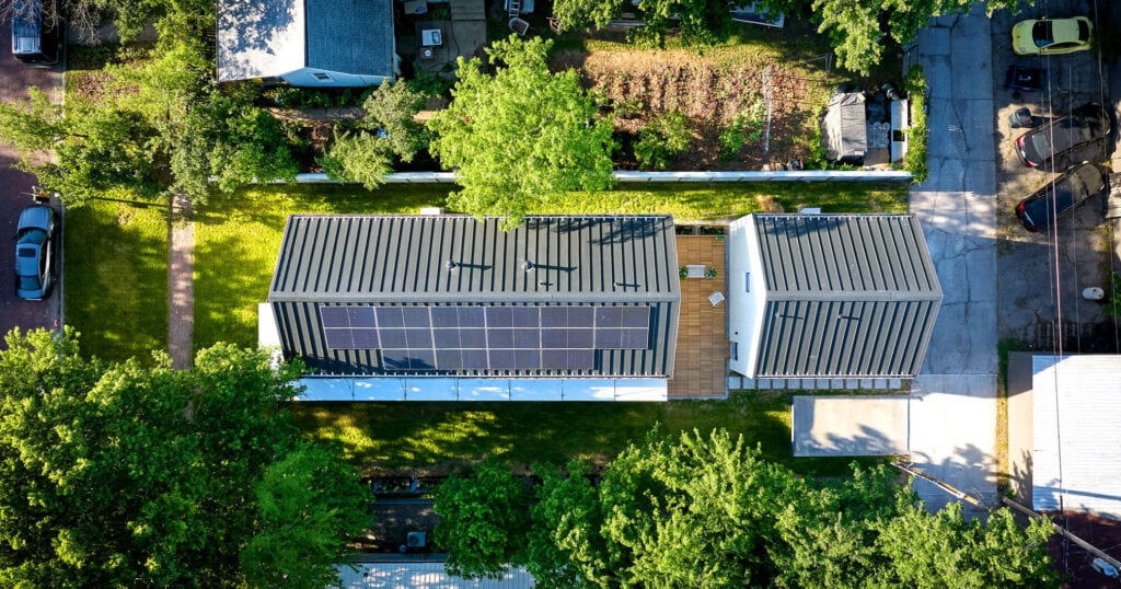 Vista aérea de una vivienda residencial moderna con techo metálico de junta alzada y paneles solares en la azotea, rodeada de árboles y casas vecinas.