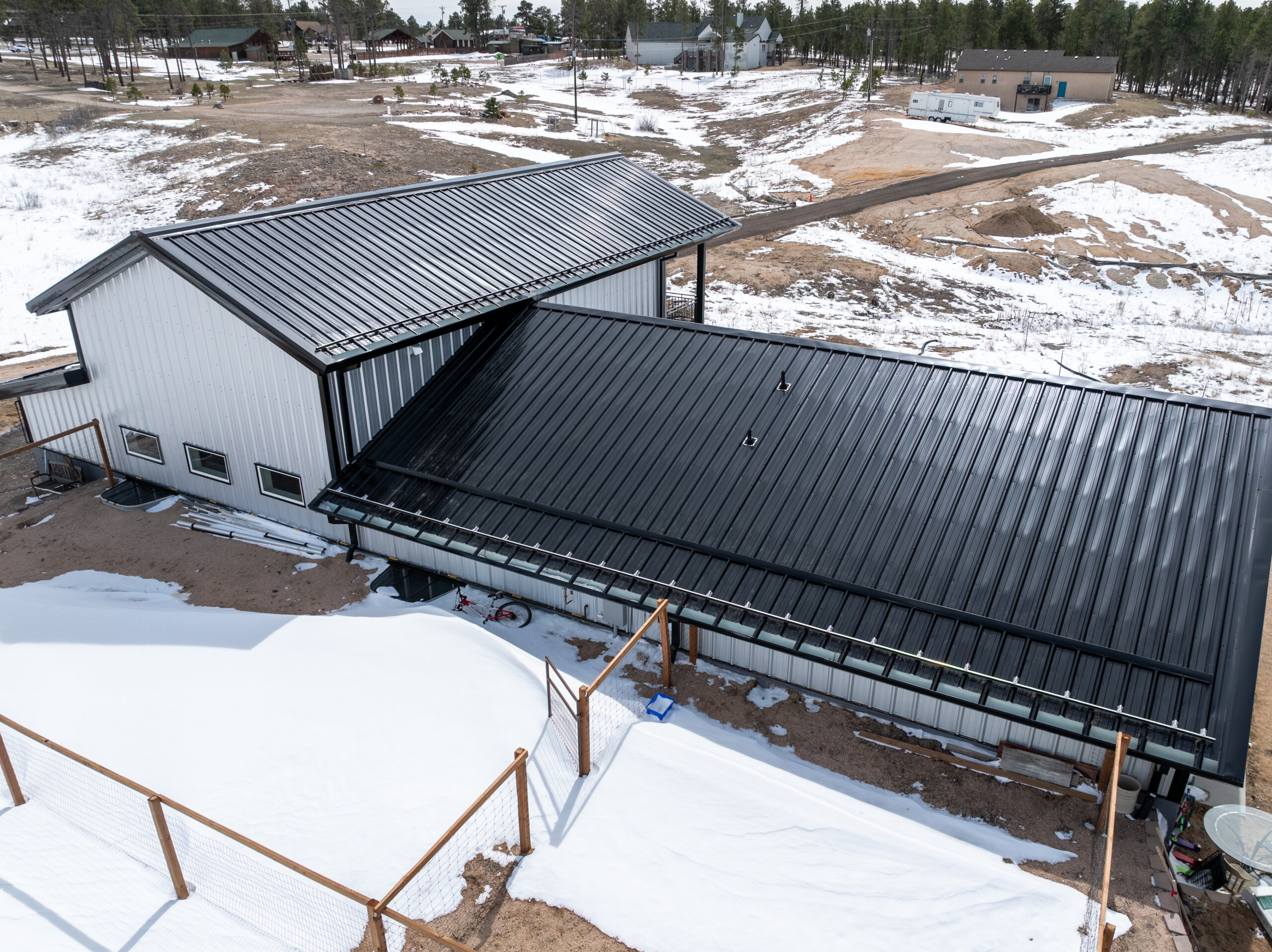 High-angle view of a grey building with a black ribbed metal roof in a snowy landscape with patches of earth.